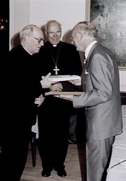 Terwijl Mgr. Niënhaus toekijkt, biedt de auteur H.W.H. Valk zijn boek "Koster in dienst van de Rooms-Katholieke Kerk" aan Mgr. Ter Schure (links) aan, 1996